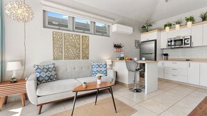 Cocina moderna con gabinetes blancos, un refrigerador de acero inoxidable y un microondas. Hay un sofá gris con cojines estampados, una mesita de madera y un taburete.