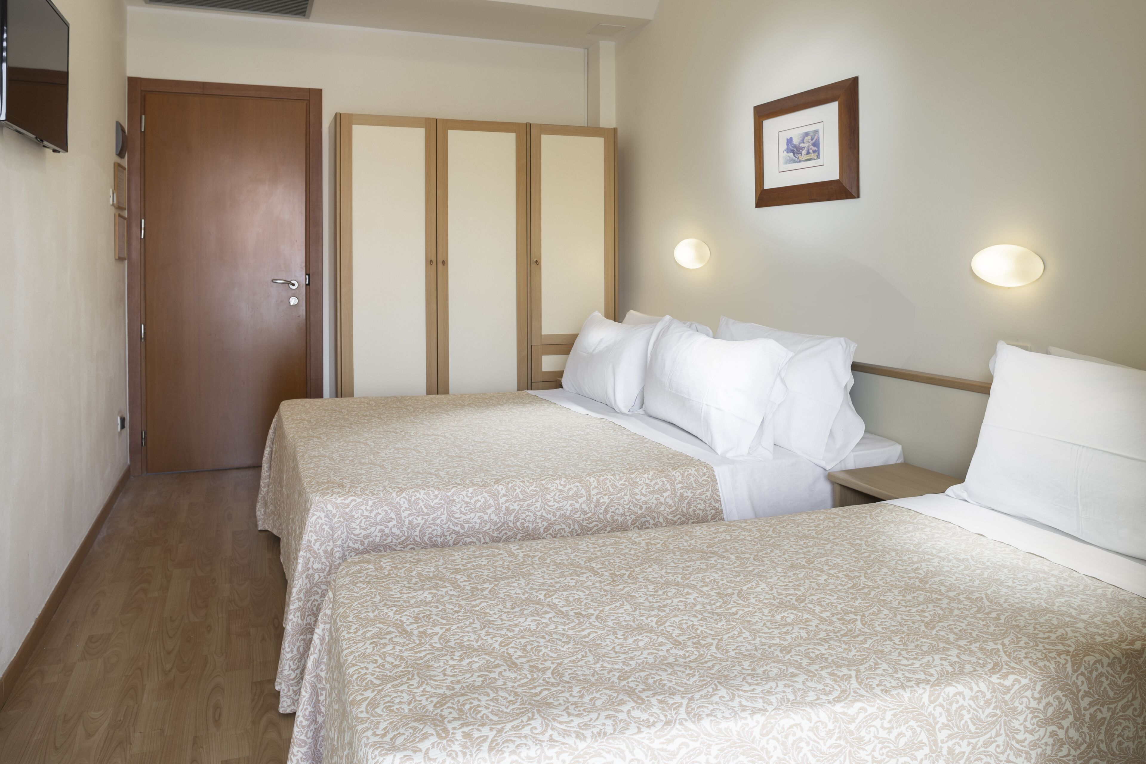 A hotel room with two beds, a wardrobe, a framed picture on the wall, and a television on the wall.
