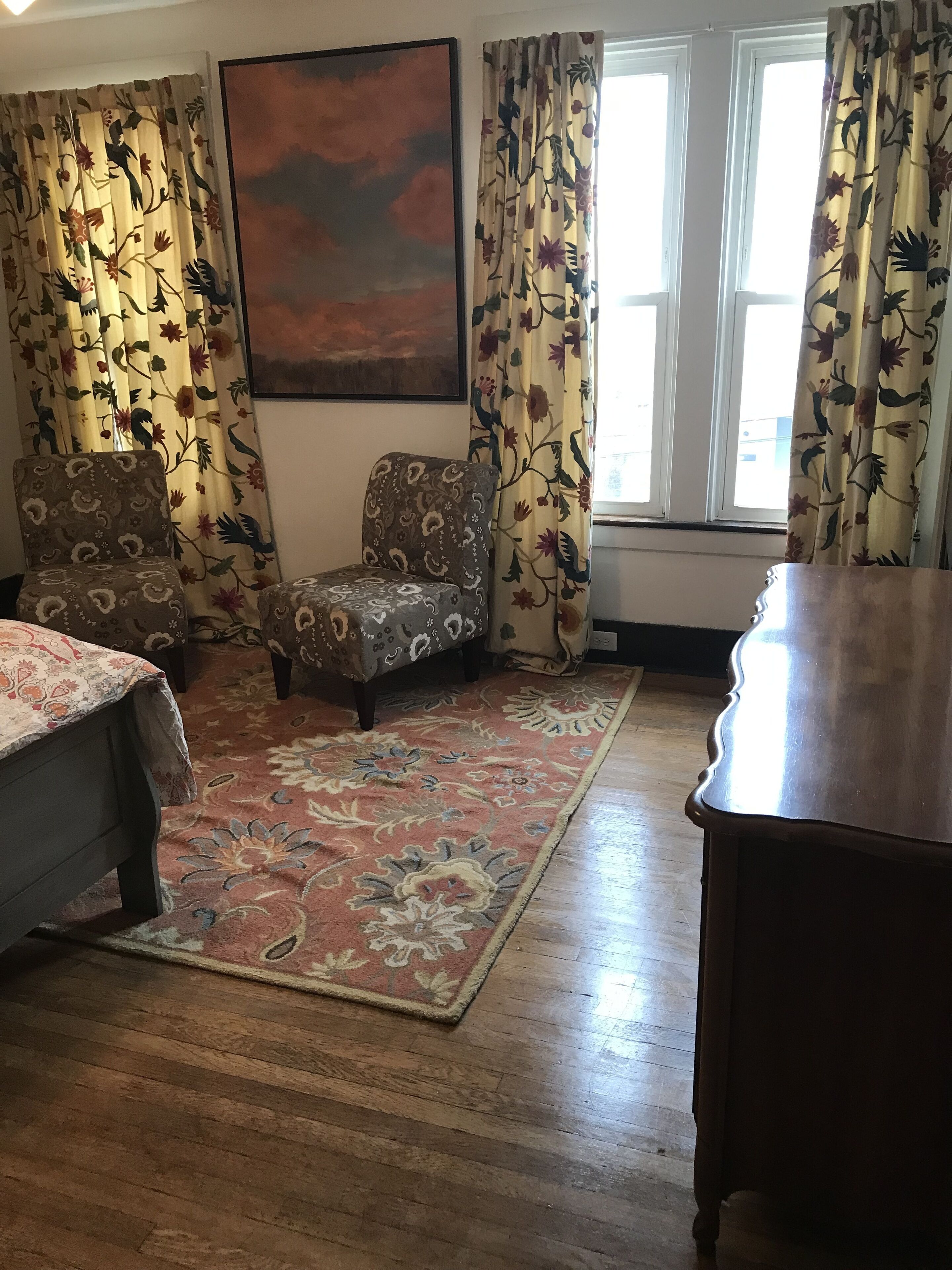 A room with a wooden floor, two patterned armchairs, a large painting on the wall, and floral curtains.