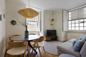 A modern living room with a glass-top dining table, wooden chairs, a sofa, a TV, and a large window with natural light. - Boutique Rentals- D*OURO Ribeira w/parking (Porto)