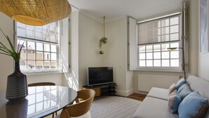 A modern living room with a glass dining table, a sofa with patterned cushions, a flat-screen TV, and large windows. - Boutique Rentals- D*OURO Ribeira w/parking (Porto)