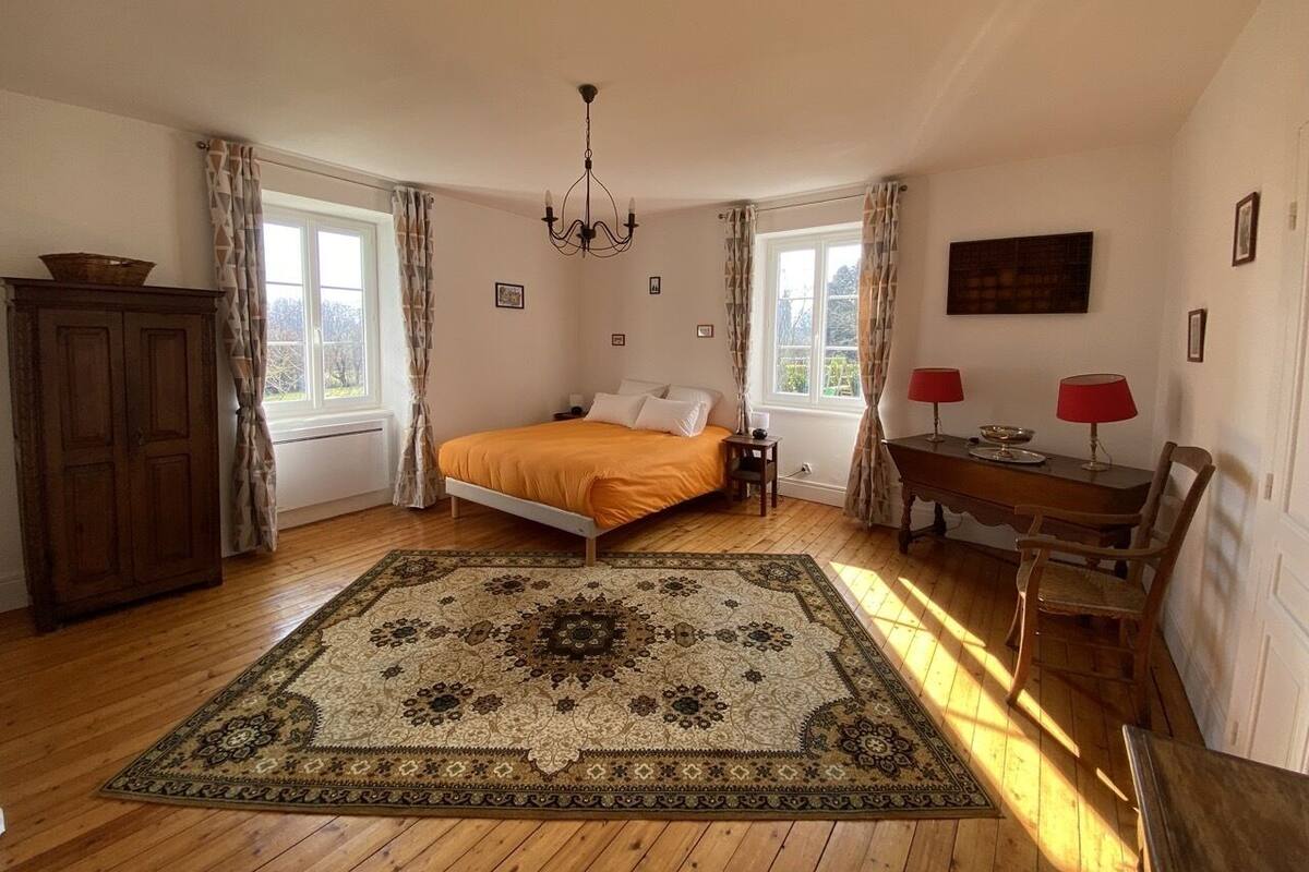 Un dormitorio con piso de madera, una cama grande con colcha naranja, un candelabro, un aparador de madera, una mesa con dos lámparas rojas y una alfombra estampada.