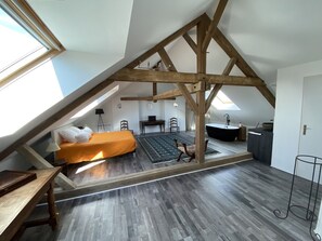 A loft bedroom with a sloped ceiling, a bed with an orange bedspread, a desk, a chair, a small table, a black chair, a black sofa, a black coffee table, a black rug, a black floor lamp, a black side table, a black cabinet, a black sink, a black countertop, and a black stool.