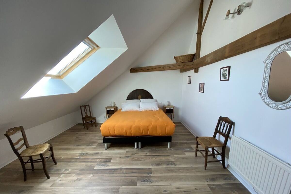 Un dormitorio con techo inclinado, un tragaluz, una cama con colcha naranja, dos sillas de madera y un radiador blanco.