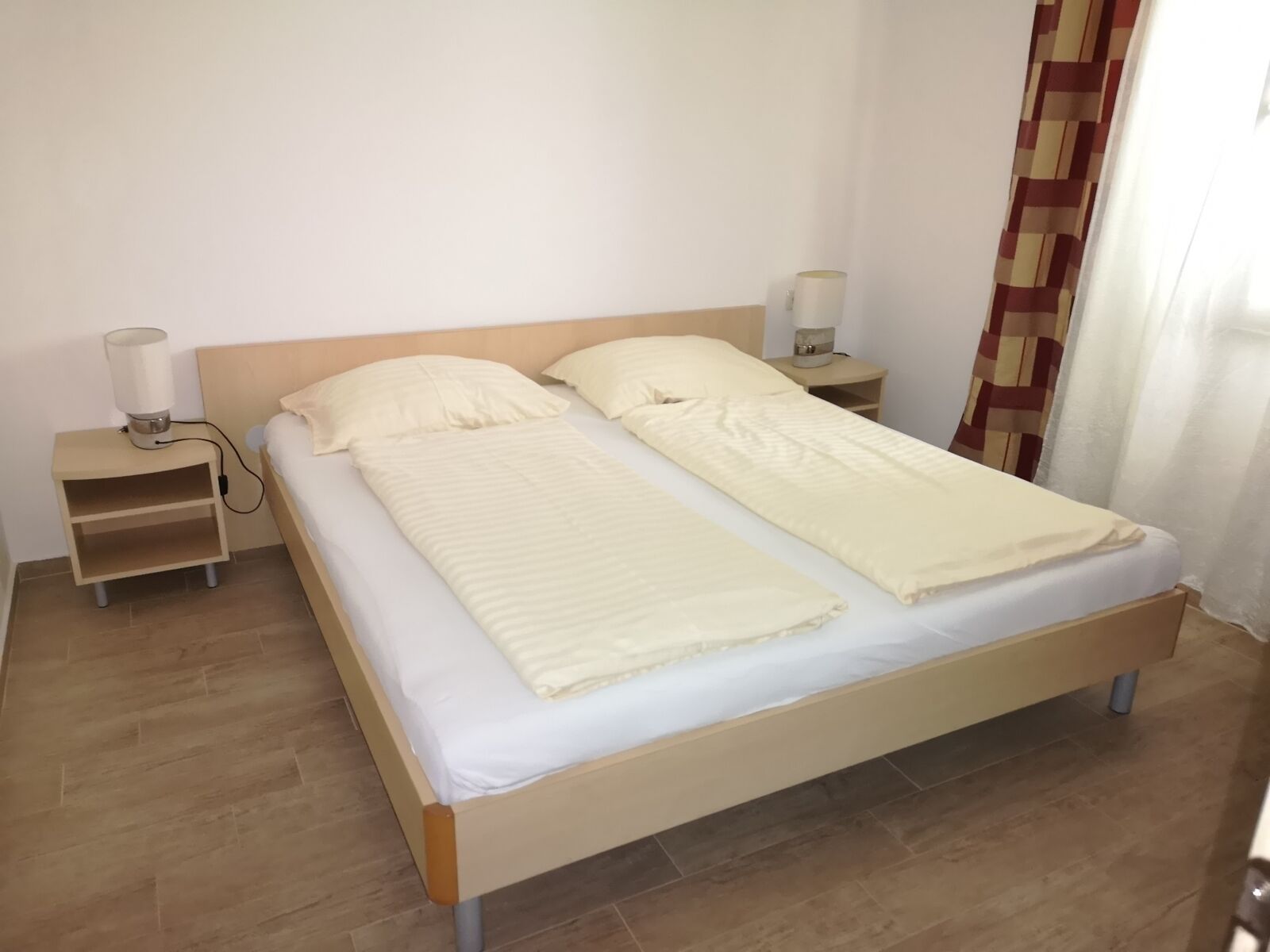 Una cama doble con estructura de madera, dos mesitas de noche, una lámpara en cada mesita y una cortina en la ventana.