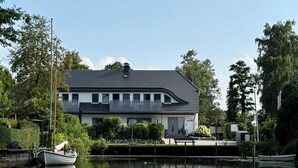 A two-story house with a balcony, a dock with a boat, and a calm water body reflecting the building.