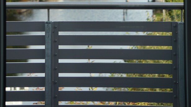 A view through a window with a metal railing, overlooking a river with trees and buildings in the background.
