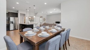 A modern kitchen with a stainless steel refrigerator, a dining area with a wooden table set for six, and a living area with a sofa and a TV.