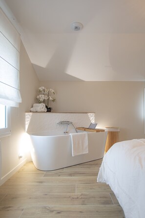 A modern bathroom with a freestanding bathtub, a vanity with a laptop, and a bedside table with a lamp.