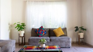 Una sala de estar con un sofá gris, cojines coloridos, dos plantas en macetas y una mesa de centro de vidrio.