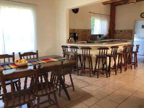 A dining area with a wooden table and chairs, a kitchen in the background, and a window with curtains. - Casa Amma with garden and pool, ideal for families and friends. (Sayulita)
