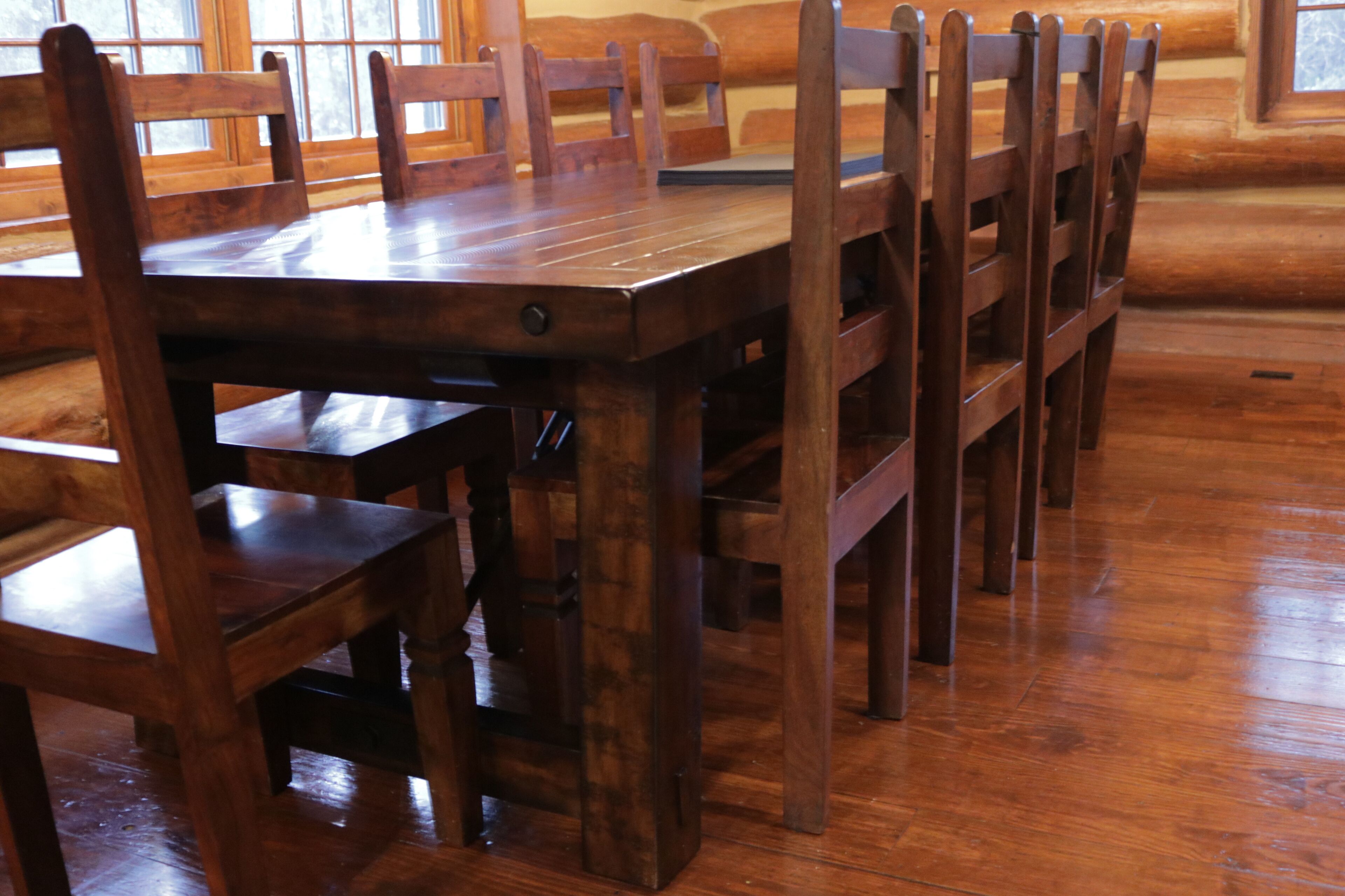 A wooden dining table with six chairs, set in a room with wooden flooring and large windows.