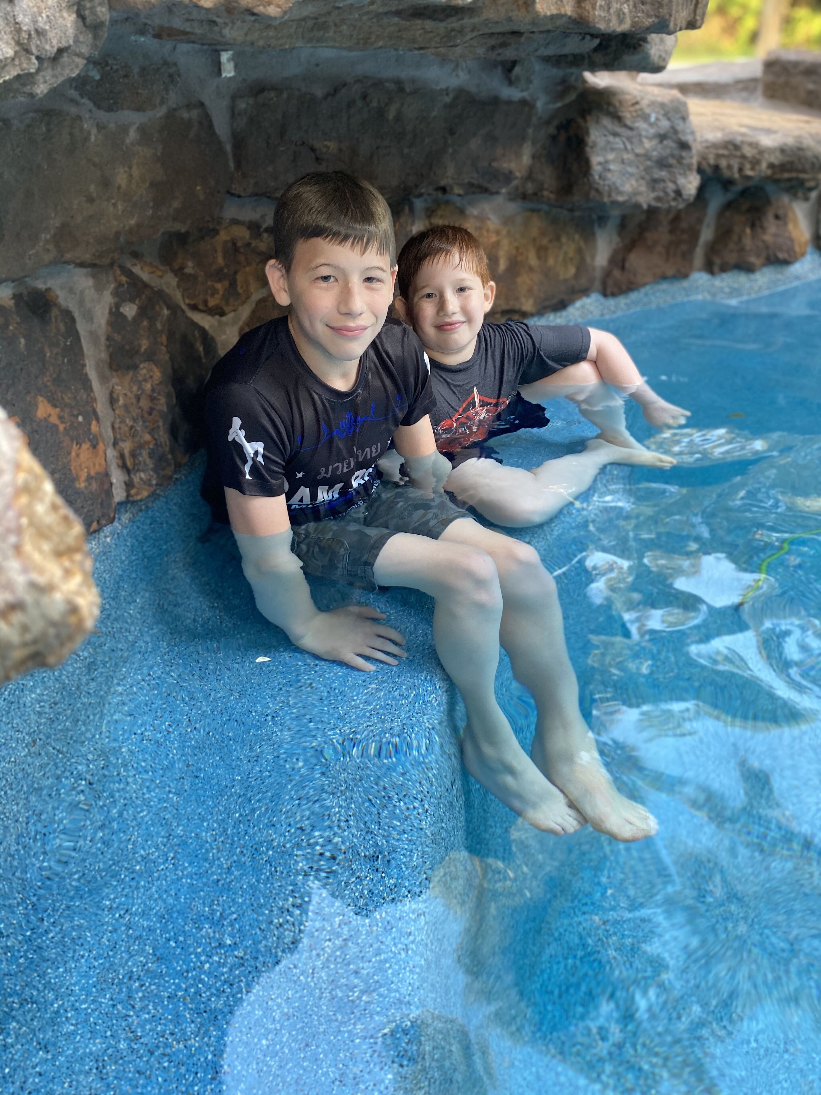 Twee kinderen zitten op een blauw oppervlak met structuur, nabij een zwembad met een rotsachtige achtergrond.