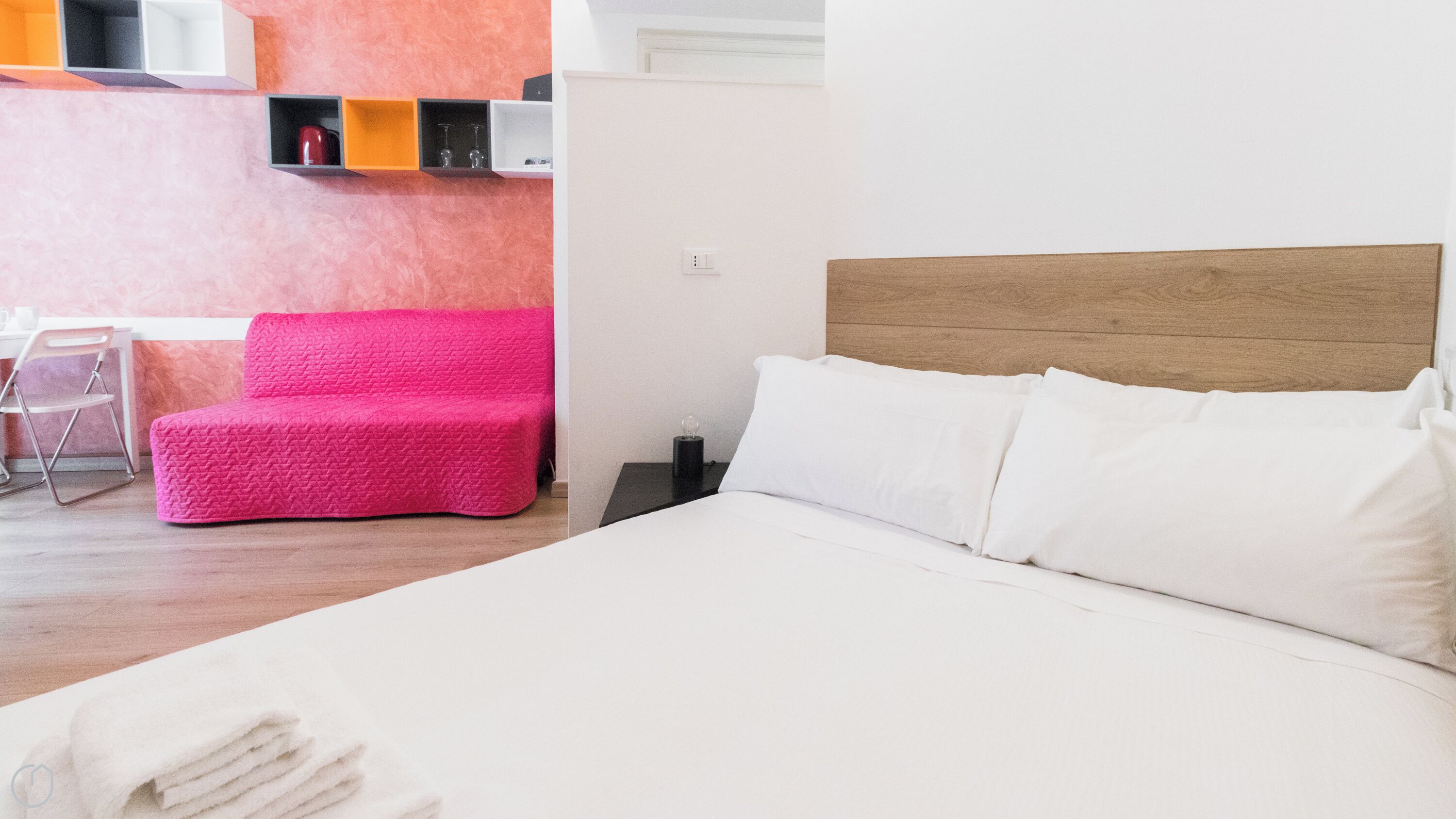 A hotel room with a bed, a chair, and a shelf with items.
