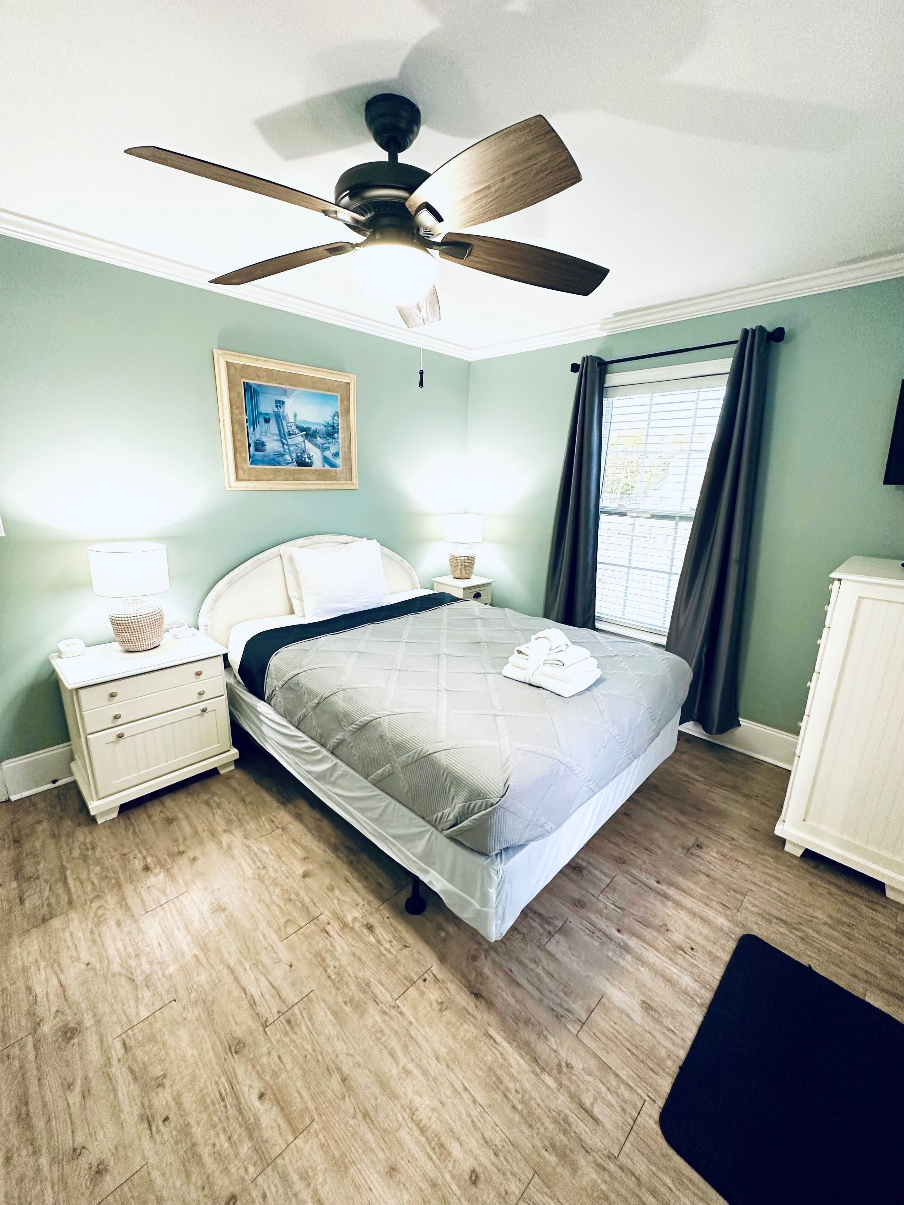 A bedroom with a bed, a nightstand, a lamp, a ceiling fan, a window with curtains, and a picture on the wall.