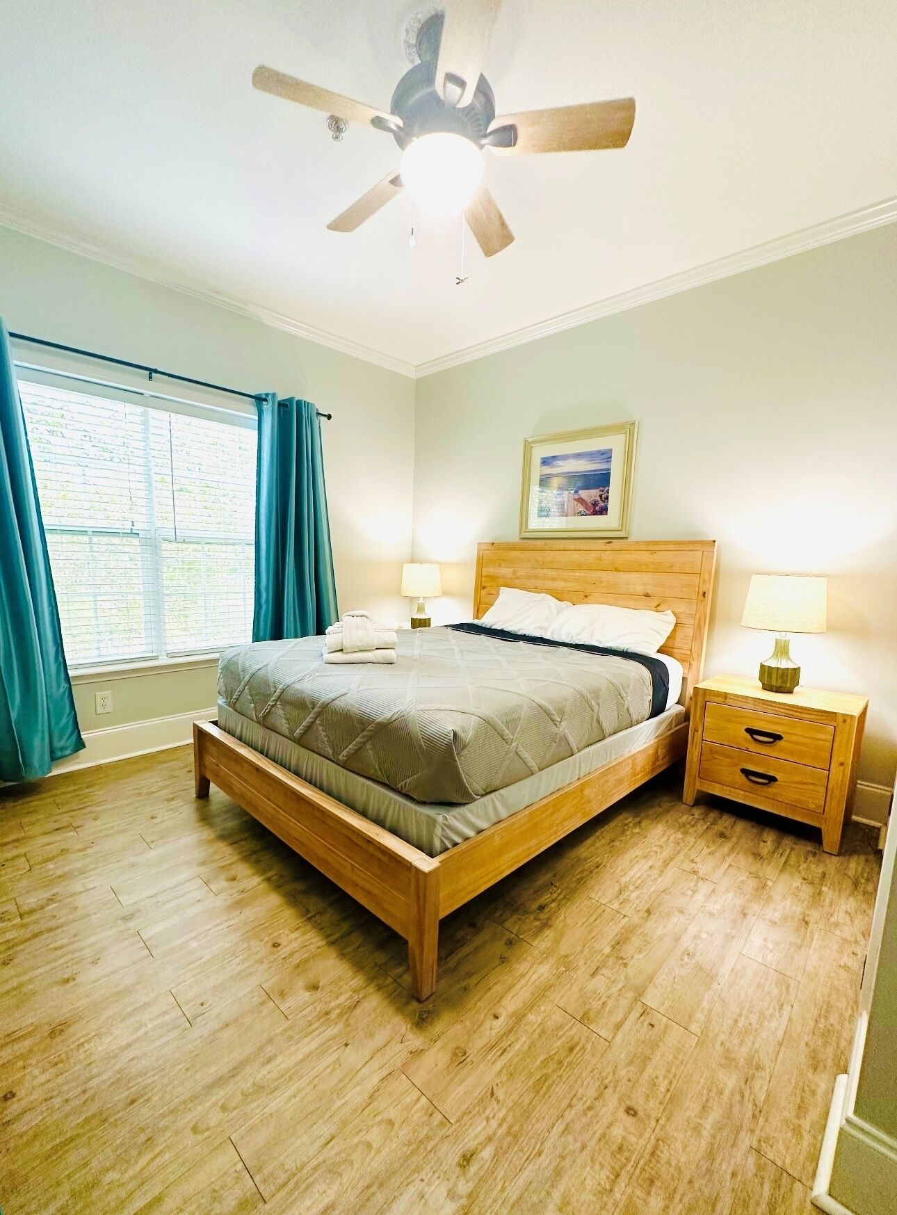 Un dormitorio con cama de madera, ventilador de techo, dos mesitas de noche con lámparas, una ventana con cortinas e imagen enmarcada en la pared.
