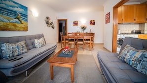 Ein Wohnzimmer mit einer Couch, einem Couchtisch und einem Essbereich mit Stühlen und Tisch. An der Wand hängt ein Gemälde, und es gibt eine Küchenzeile mit Schränken und Geräten.