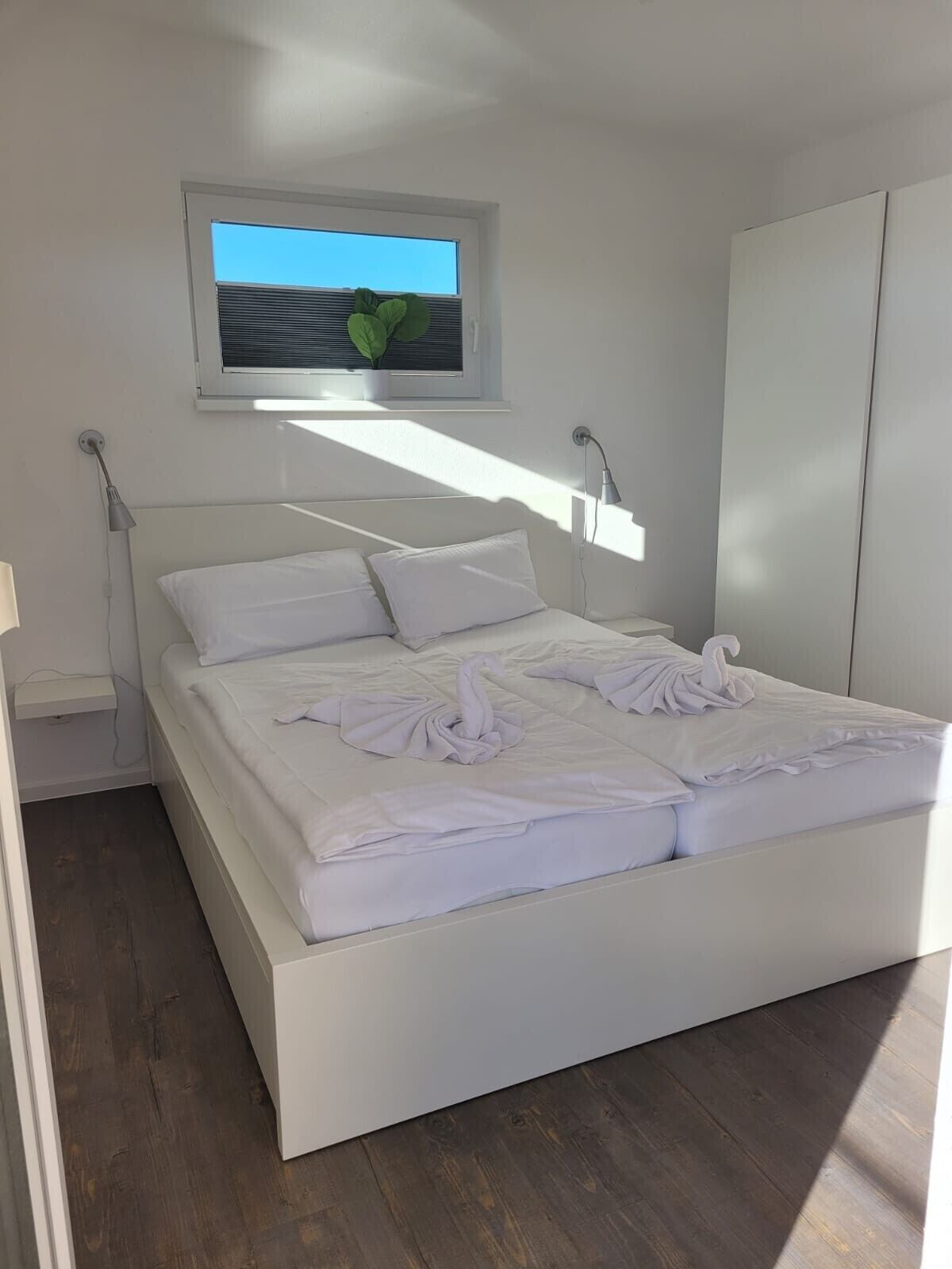 A modern bedroom with a large bed, a window with blinds, a bedside lamp, and a wardrobe.