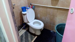A small bathroom with a toilet, a trash bin, and a shelf with cleaning supplies. - Indo Hometown (Garut)