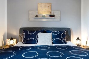 Un lit recouvert d’une couvre-lit à motifs bleus et blancs, un oreiller blanc et des coussins assortis. Au-dessus du lit, on trouve une étagère sur laquelle sont posés un petit modèle de bateau et un tableau.