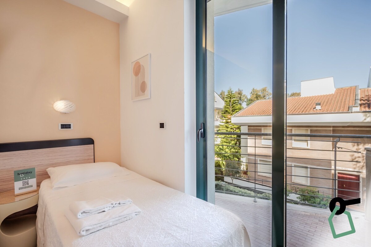 Hotelkamer met een bed, nachtkastje, een wandlamp en balkon met uitzicht op gebouwen en bomen.