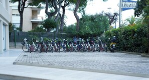 Uma fileira de bicicletas estacionadas em área pavimentada, com um edifício e árvores ao fundo.