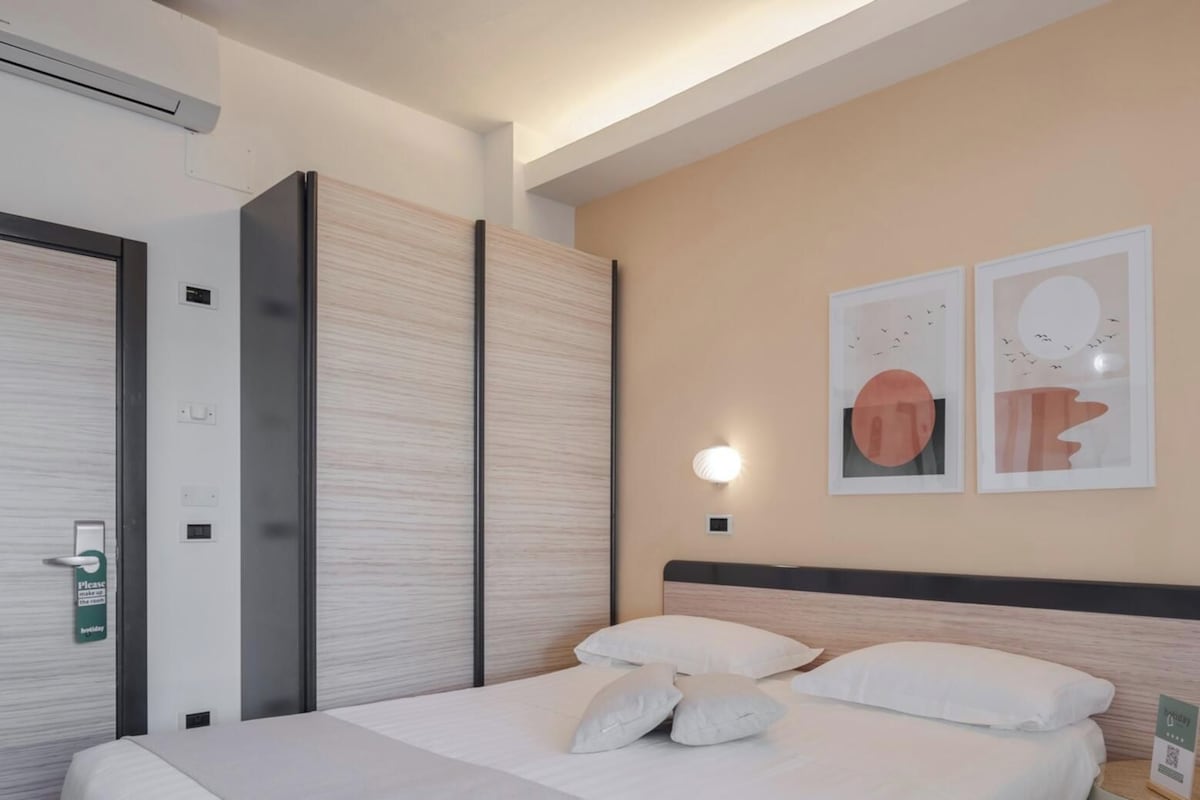 Une chambre d’hôtel avec un lit, des oreillers, une lampe fixée au mur et des œuvres d’art.