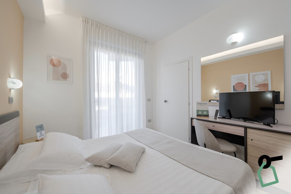 Une chambre d’hôtel avec un lit, un bureau avec une télévision, une chaise et une fenêtre avec des rideaux.