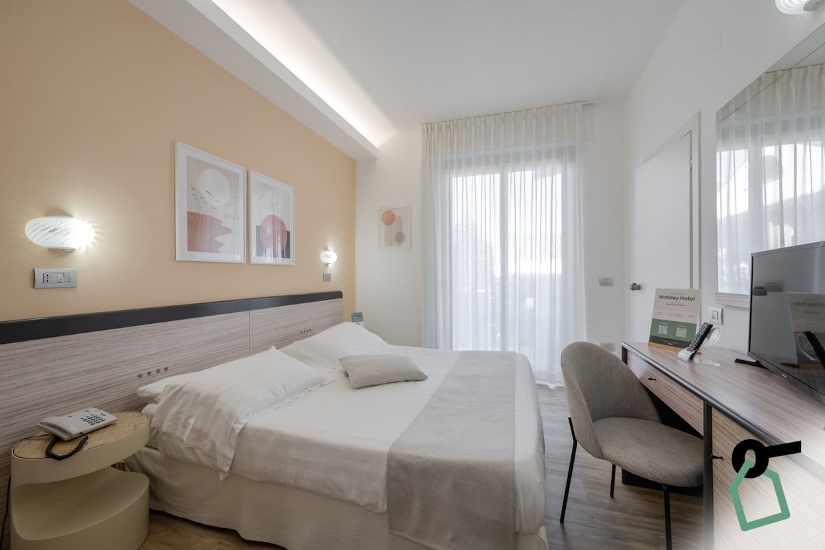 Quarto de hotel com cama, escrivaninha, cadeira, televisão e janela com cortinas.