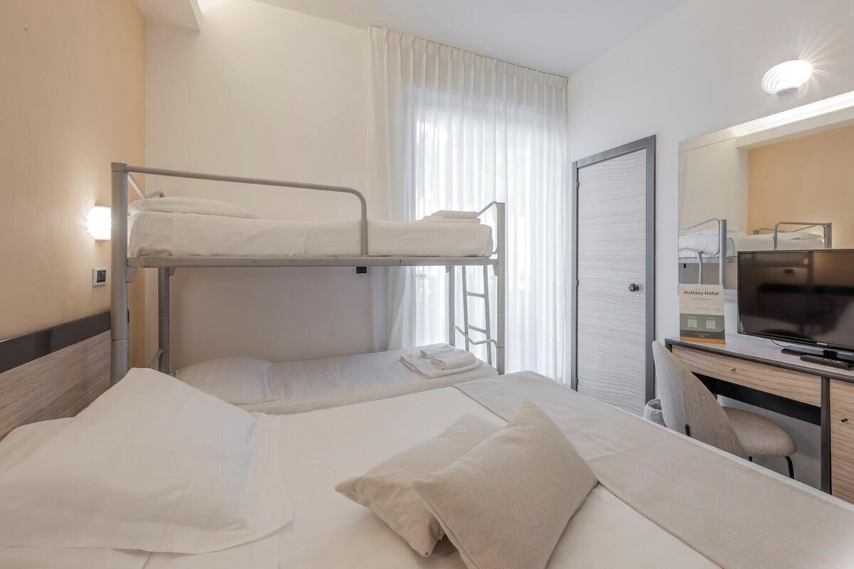 Quarto de hotel com beliches, escrivaninha com cadeira, televisão e porta do banheiro.
