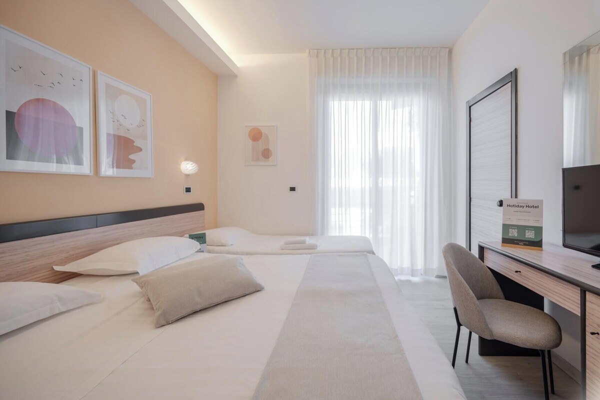Quarto de hotel com cama, escrivaninha, cadeira e televisão.