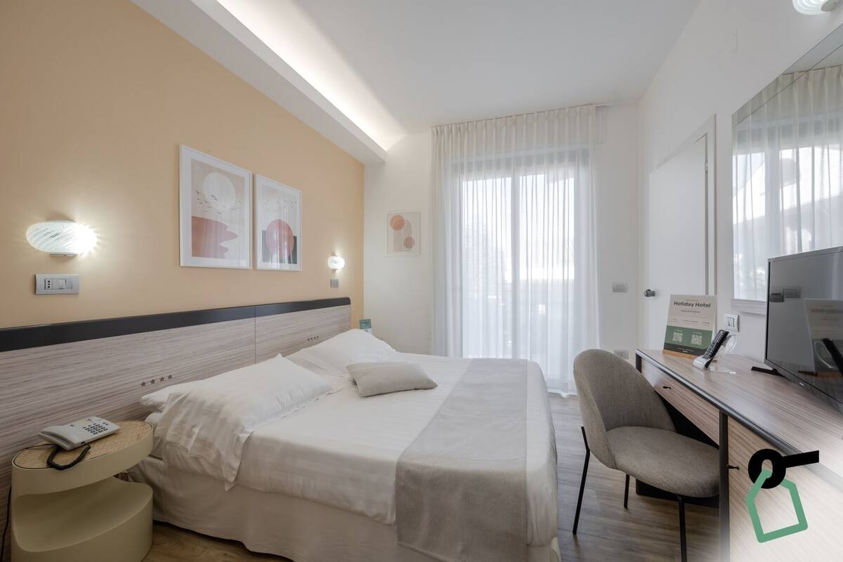 Een hotelkamer met een bed, nachtkastje met telefoon, bureau met stoel, televisie en raam met gordijnen.
