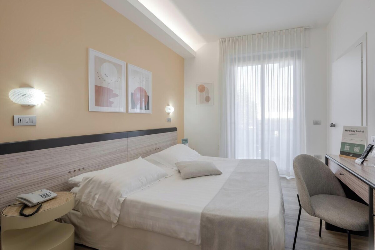 Quarto de hotel com cama, mesa de cabeceira, abajur e escrivaninha com cadeira.
