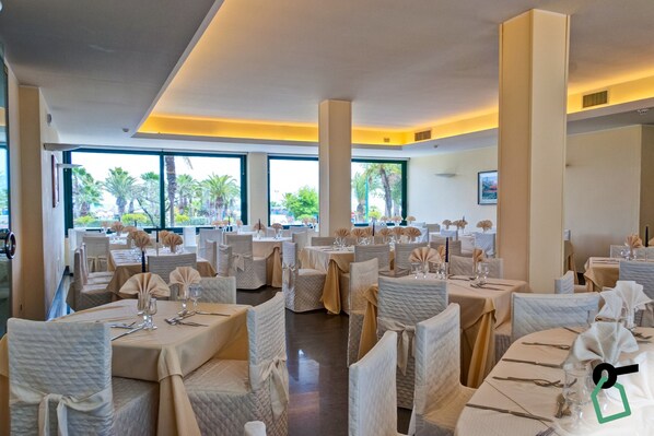 Une salle à manger spacieuse, avec des tables rondes, des chaises blanches et de grandes fenêtres donnant sur des palmiers.