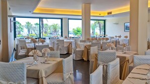 Een restaurant met meerdere eettafels, grote ramen met uitzicht op palmbomen en een modern interieur.