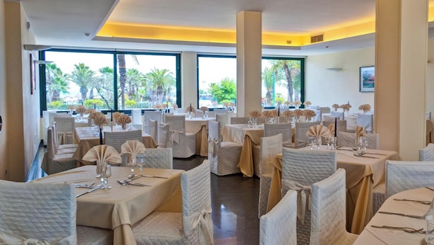 Un restaurante con varias mesas dispuestas para comer, amplias ventanas con vista a palmeras y una decoración interior moderna.