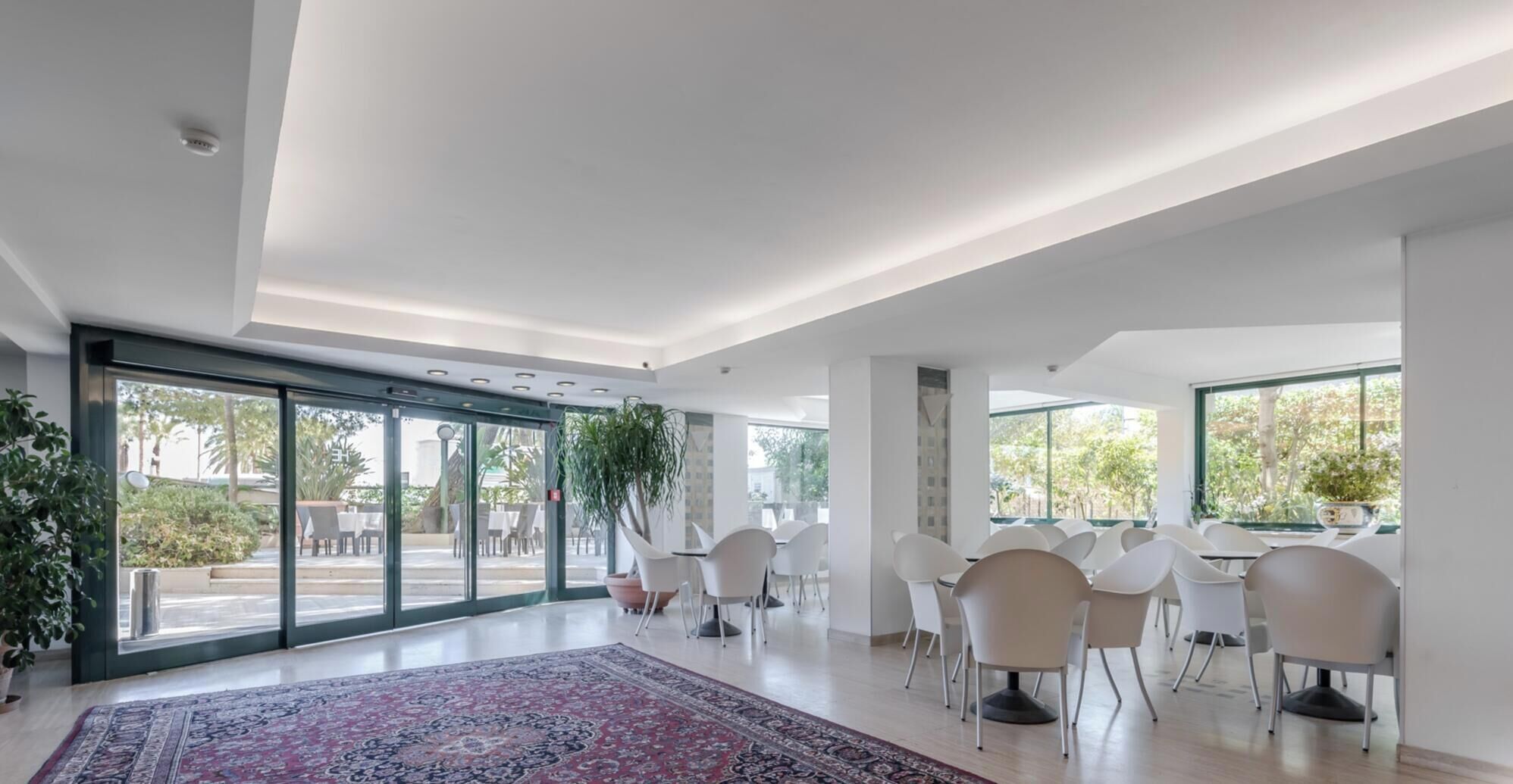 Una habitación amplia con un gran ventanal corredero que da a un espacio exterior, sillas blancas dispuestas para sentarse y una alfombra estampada en el suelo.