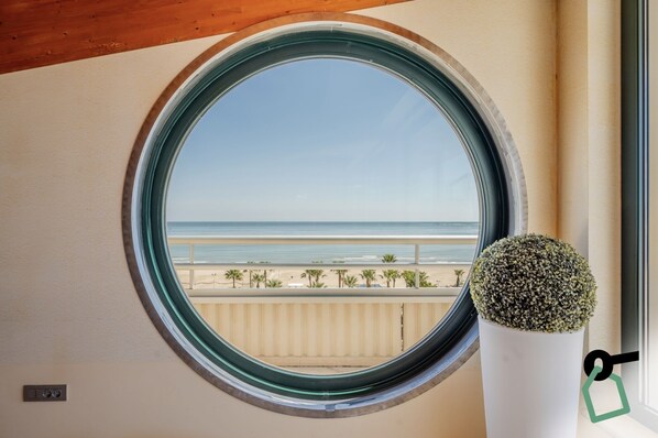 Janela redonda com vista para a praia e palmeiras.