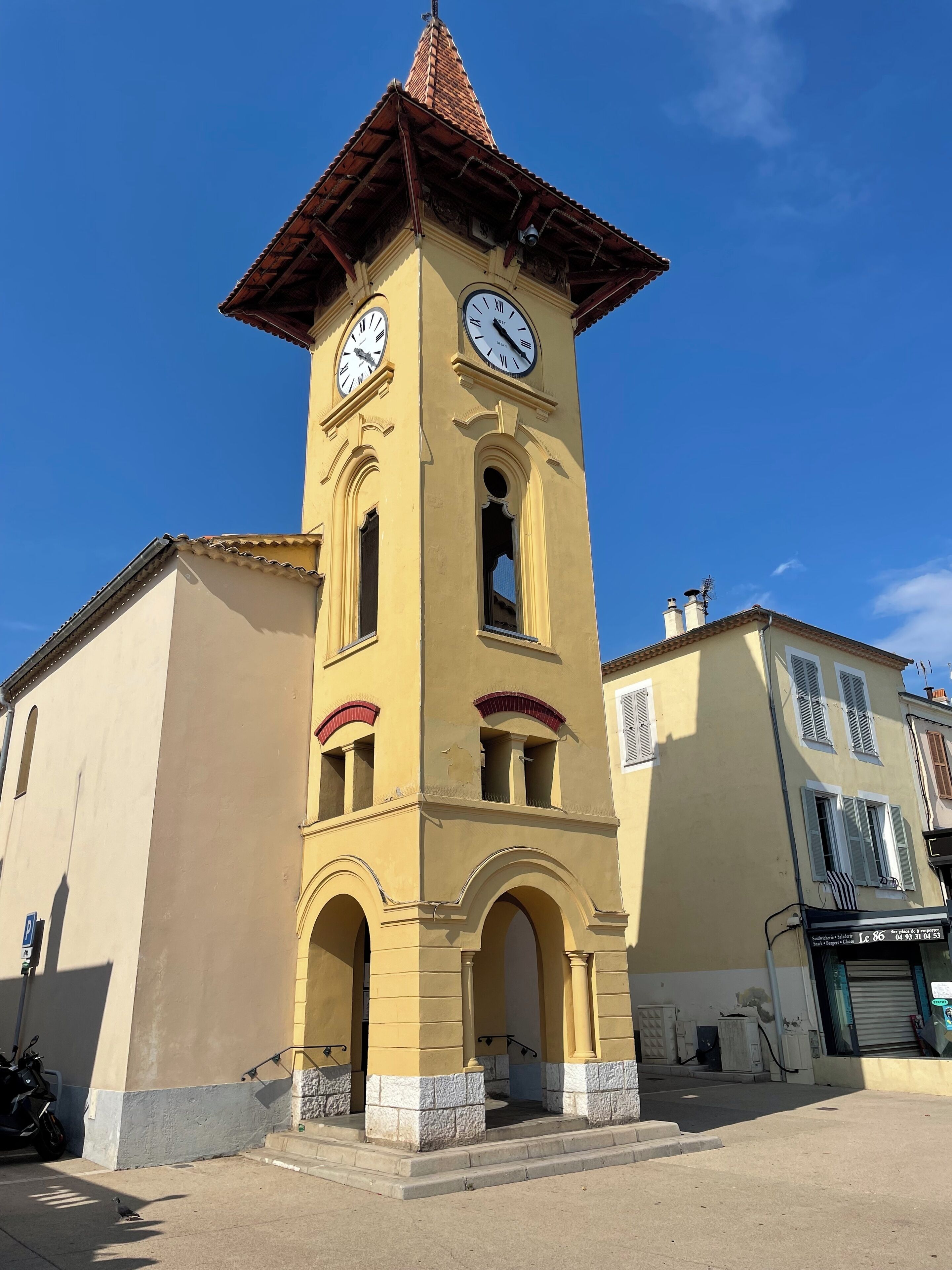 Une tour jaune, dotée d’une horloge, de fenêtres en arc de cercle et d’un toit recouvert de tuiles.