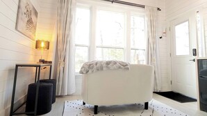 Une pièce avec une chaise blanche, une table avec une lampe et une fenêtre avec des rideaux.