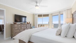 Un dormitorio con una cama grande, un ventilador de techo, una televisión, un aparador de madera y vistas al océano a través de amplias ventanas.