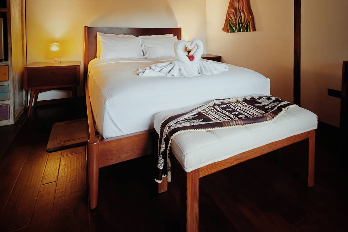 Una cama con estructura de madera, una toalla con forma de corazón, una lámpara de noche y una pintura en la pared.
