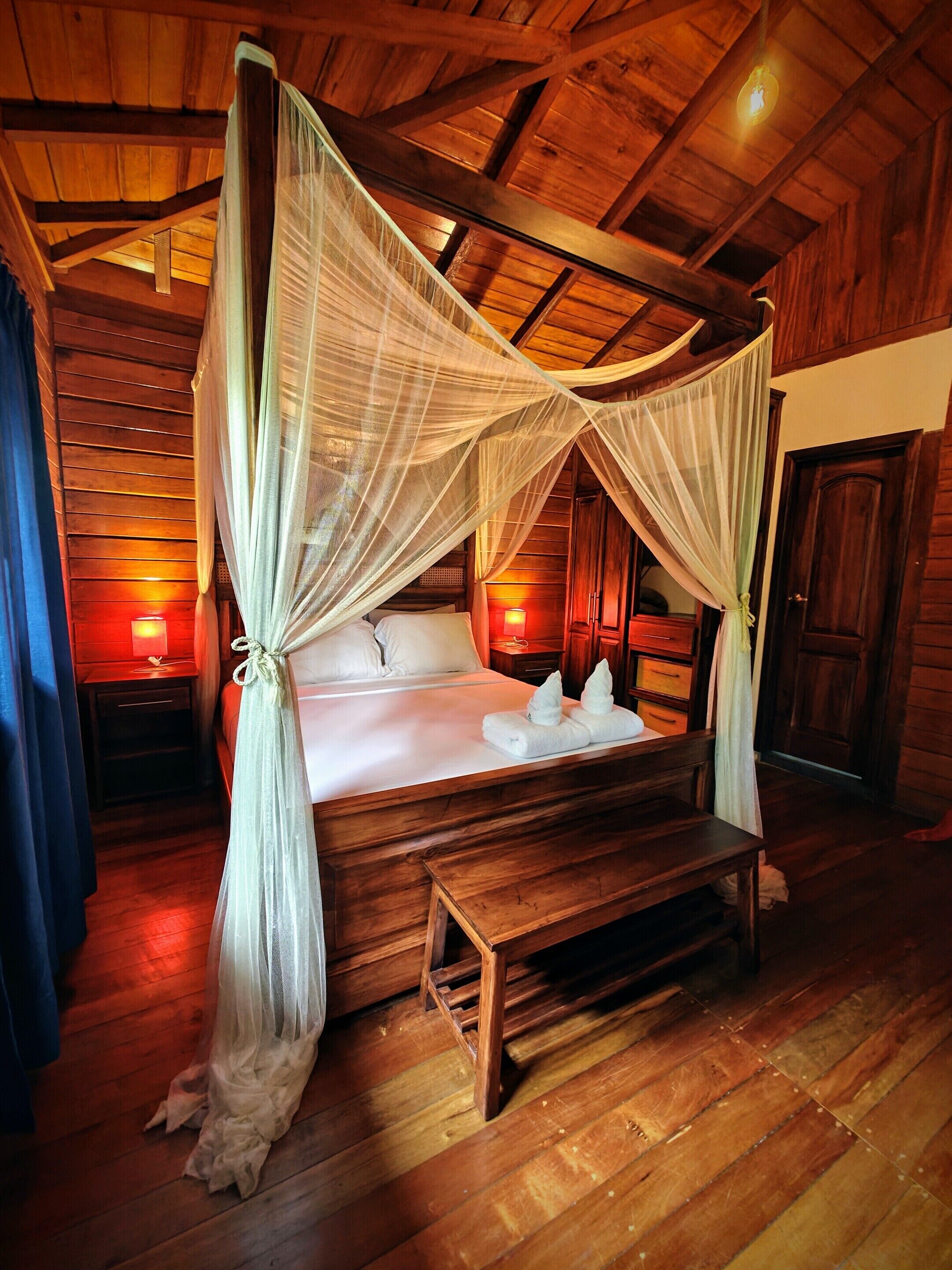 Uma cama de madeira com dossel, abajures laterais e um banquinho.