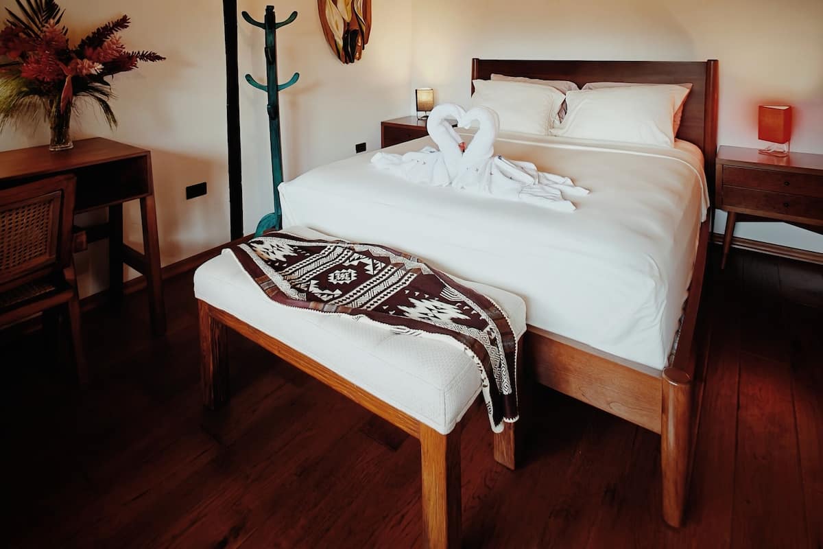 Un dormitorio con techo de madera, una cama con ropa de cama blanca, un banco, una mesita de noche, una lámpara y un adorno en la pared.