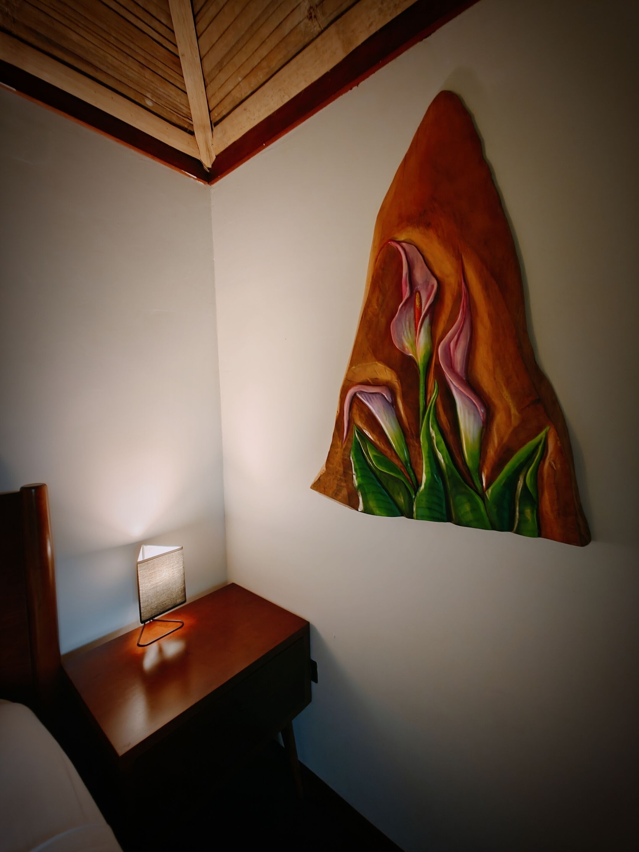 Una habitación con un escritorio de madera, una lámpara y un cuadro floral en la pared.