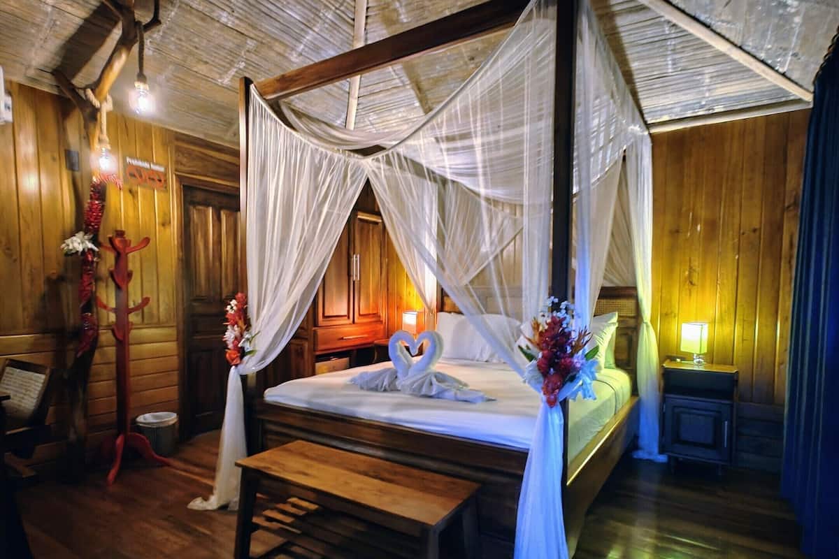 Una cama con dosel sostenido por cuatro pilares, suelo de madera, un banco y una mesita de noche con lámpara.