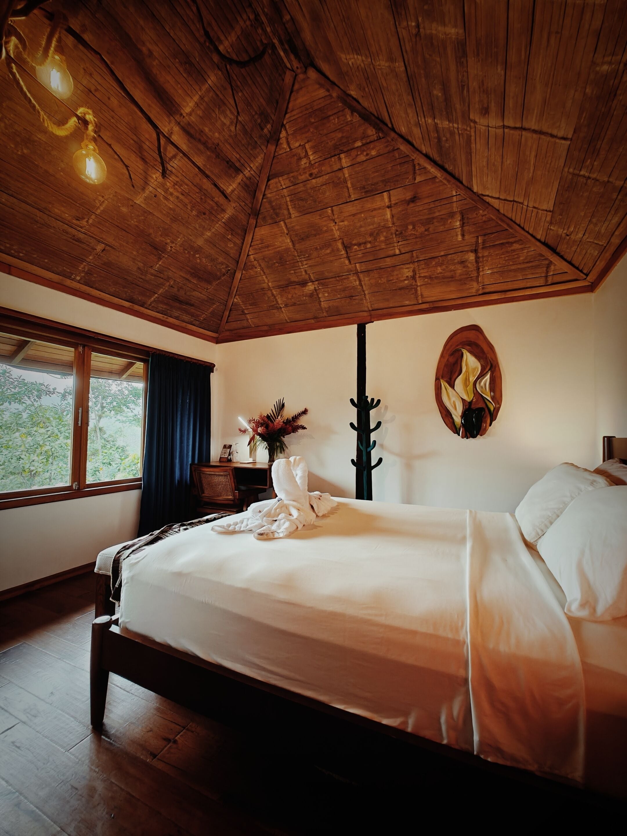 A bedroom with a wooden ceiling, a bed with white linens, a window with curtains, and a wall-mounted artwork.