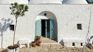 A white building with a central archway and a door, surrounded by potted plants and a small tree.