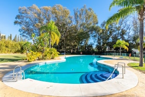 A swimming pool with a curved design, surrounded by a paved deck and palm trees. - Bahia de Marbella | Bright Beachside Apartment with Pool in Marbella (Marbella)