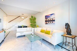 A modern living room with a white sofa, a glass coffee table, and a staircase leading to an upper floor. - Bahia de Marbella | Bright Beachside Apartment with Pool in Marbella (Marbella)